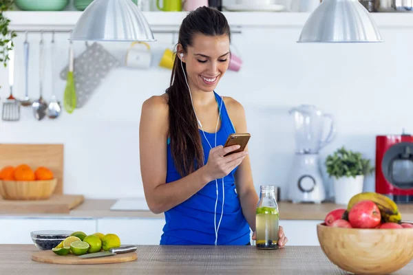 Jovem esportiva ouvindo música com telefone celular depois de treinar na cozinha em casa . — Fotografia de Stock