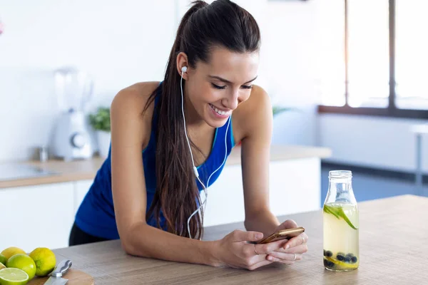 Mujer joven deportiva escuchando música con teléfono móvil después de entrenar en la cocina en casa . — Foto de Stock