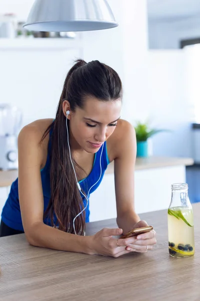 Mujer joven deportiva escuchando música con teléfono móvil después de entrenar en la cocina en casa . —  Fotos de Stock