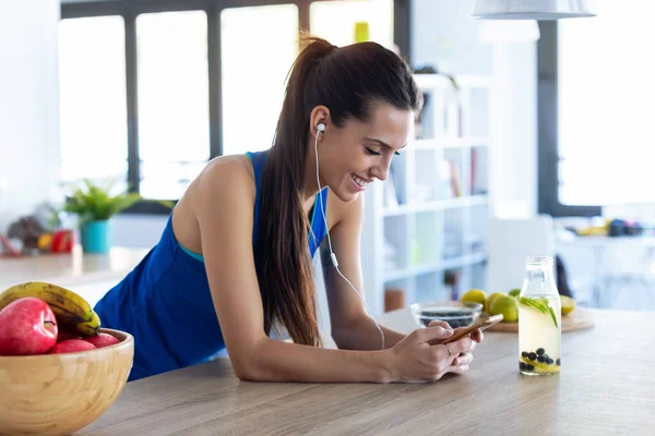 Sportovní mladá žena poslech hudby s mobilním telefonem po tréninku v kuchyni doma. — Stock fotografie