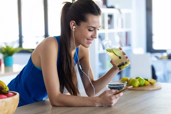 Sportowa młoda kobieta słuchająca muzyki z telefonu komórkowego po treningu w kuchni w domu. — Zdjęcie stockowe