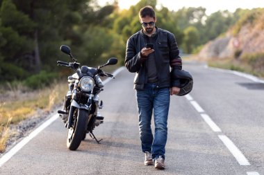 Yolda bir arıza yaşadıktan sonra yolda yürürken cep telefonunu kullanan bir erkek motorcu..