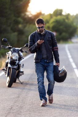 Yolda bir arıza yaşadıktan sonra yolda yürürken cep telefonunu kullanan bir erkek motorcu..