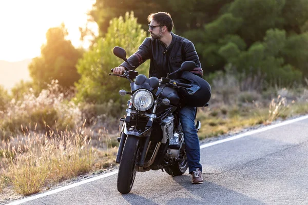 Beau motard barbu regardant sur le côté tout en étant assis sur sa moto et tenant un casque . — Photo