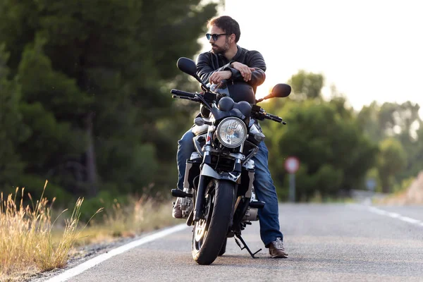 Beau motard barbu regardant sur le côté tout en étant assis sur sa moto et tenant un casque . — Photo