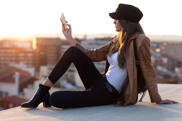 Hübsche junge Frau macht ein Selfie mit Handy, während sie auf dem Dach sitzt. — Stockfoto