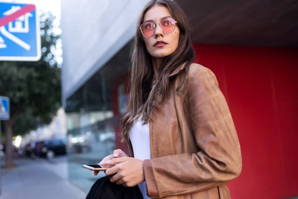 Mujer bastante joven mirando hacia los lados mientras usa su teléfono móvil en la calle . — Foto de Stock