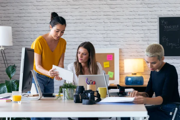 Drie moderne zakenvrouwen praten en evalueren het laatste werk op de digitale tablet in een gezamenlijke werkruimte. — Stockfoto