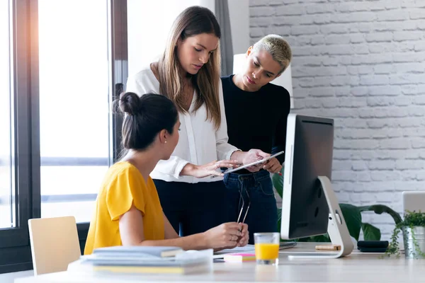 Tre moderna affärskvinnor pratar och granskar det senaste arbetet med den digitala surfplattan på en gemensam arbetsplats. — Stockfoto