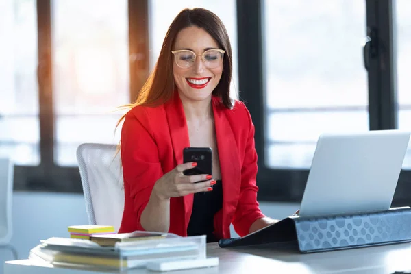 Zâmbind tânără femeie de afaceri folosind telefonul mobil în timp ce lucra cu computerul la birou . — Fotografie, imagine de stoc