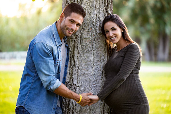 Pretty pregnant couple looking at the camera while holding hands in the park.