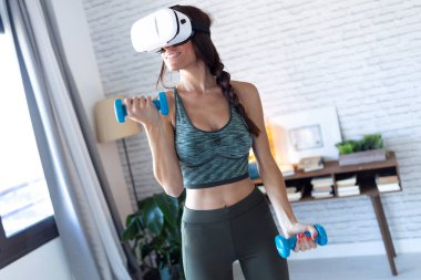 Güzel, genç bir kadın, evdeki sanal gerçeklik kulaklığıyla dambıllarla egzersiz yapıyor..