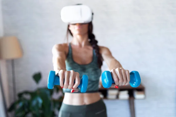 Hermosa joven haciendo un entrenamiento con pesas con auriculares de realidad virtual en casa . —  Fotos de Stock