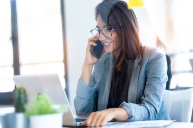 Ofiste dizüstü bilgisayarıyla çalışırken gülümseyen bir iş kadınının cep telefonuyla konuşması..