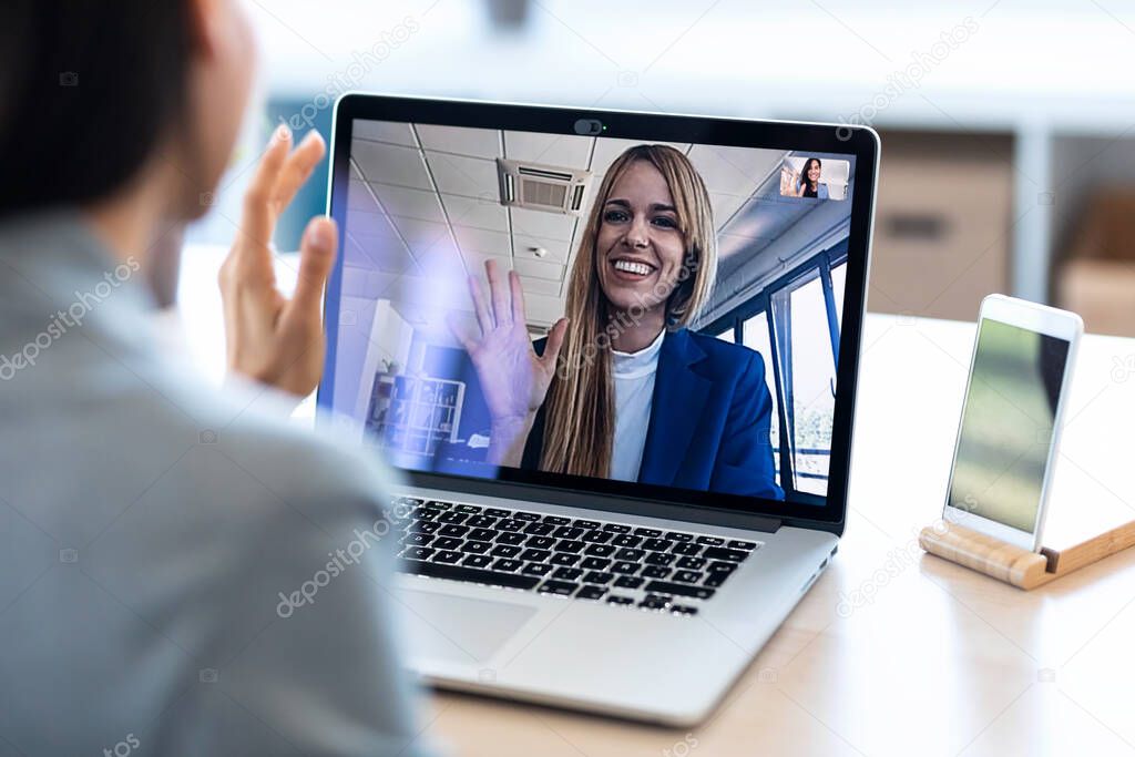 Back view of female employee waving and speaking on video call with her colleague on online briefing with laptop at home.