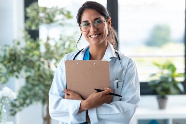 Kameraya bakıp gülümseyen, elinde tıbbi belgeler olan ve muayenede ayakta duran güzel bir kadın doktor fotoğrafı..