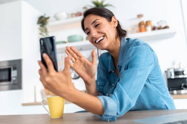 Güzel bir genç kadının akıllı telefonuyla video görüşmesi yaparken ve evde sabah kahvesini içerken el sallarken..