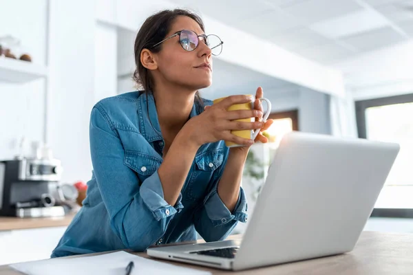 自宅のキッチンでラップトップで作業しながら かなり若い女性のショットは 1つの瞬間をリラックスしてコーヒーを飲む — ストック写真