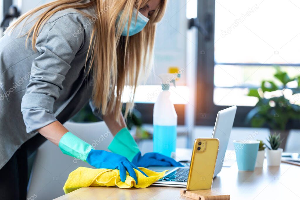 Close-up of young business woman with face mask and protective gloves cleaning the laptop with spray and rag in office.