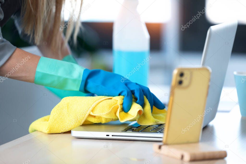 Close-up of woman with protective gloves cleaning the laptop with spray and rag in office.