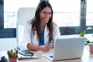 Konsültasyondaki bir bilgisayarla iş arkadaşlarıyla görüntülü konuşma yapan kadın doktor..