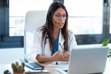 Konsültasyondaki bir bilgisayarla iş arkadaşlarıyla görüntülü konuşma yapan kadın doktor..