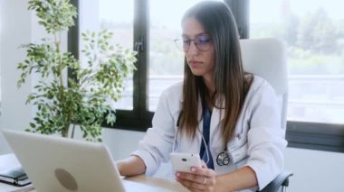 Danışmanlıkta dizüstü bilgisayar üzerinde çalışırken cep telefonunu kullanan kendine güvenen genç bir kadın doktorun videosu..