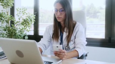 Danışmanlıkta dizüstü bilgisayar üzerinde çalışırken cep telefonunu kullanan kendine güvenen genç bir kadın doktorun videosu..