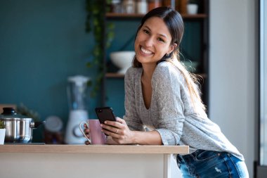 Gülümseyen genç bir kadının cep telefonunu kullanırken kameraya bakıp evdeki mutfakta bir fincan kahve içerken çekilmesi..