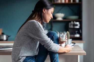 Evdeki mutfakta yoğurt yerken cep telefonunu kullanan konsantre genç bir kadın..