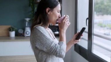 Gülümseyen genç bir kadının evdeki mutfakta kahve içerken cep telefonunu kullanmasının videosu..