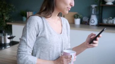 Gülümseyen genç bir kadının evdeki mutfakta kahve içerken cep telefonunu kullanmasının videosu..