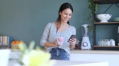 Gülümseyen genç bir kadının evdeki mutfakta kahve içerken cep telefonunu kullanmasının videosu..