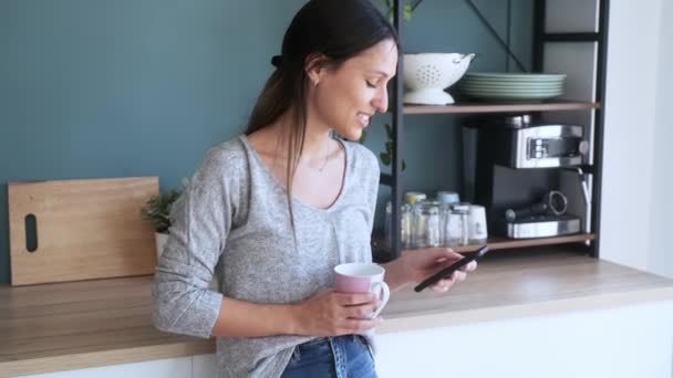 自宅のキッチンでコーヒーを飲みながら携帯電話を使って笑顔の若い女性のビデオ — ストック動画