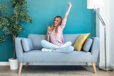 Başarılı bir genç kadının cep telefonunu kullanması ve evdeki koltukta otururken bir şeyleri kutlaması..