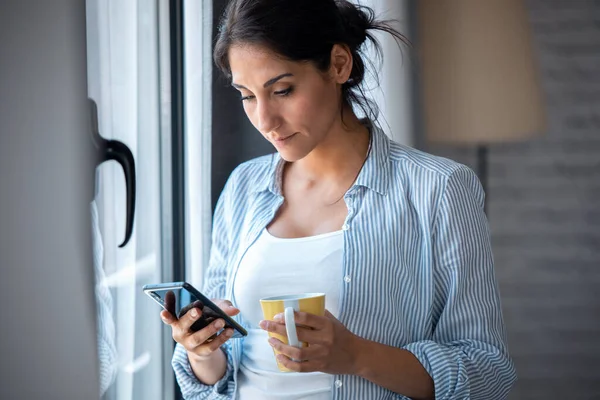 自宅のリビングルームの窓の近くにコーヒーを飲みながら彼女の携帯電話を使用してかなり若い女性のショット — ストック写真