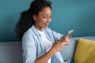 Evdeki kanepede otururken gülümseyen Afrikalı Amerikalı genç bir kadının cep telefonunu kullanması..