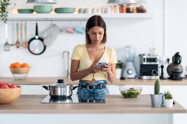 Evde, mutfakta salata yerken cep telefonunu kullanan konsantre genç bir kadının fotoğrafı..