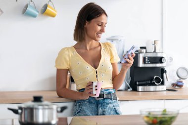 Evdeki mutfakta bir fincan kahve içerken genç ve güzel bir kadının cep telefonunu kullanması..