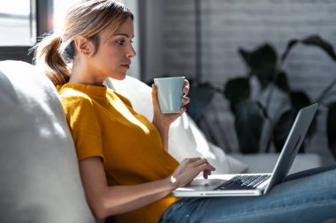 Evdeki bir kanepede oturup bir fincan kahve içerken dizüstü bilgisayarıyla çalışan kendine güvenen genç bir kadının fotoğrafı.