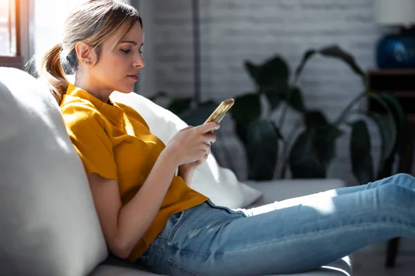 自信に満ちた若い女性のショット自宅でソファに座って携帯電話を使用して — ストック写真