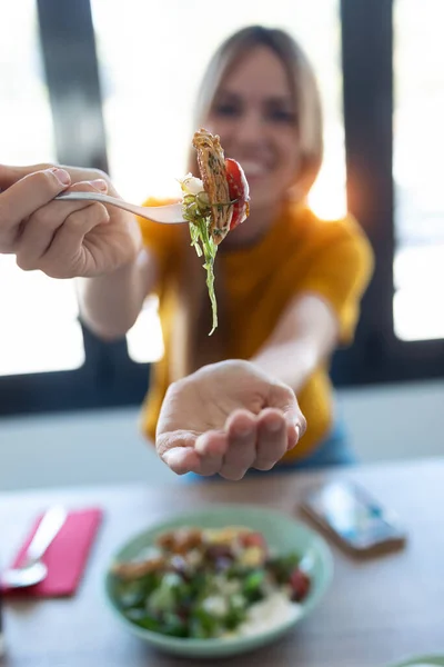自宅で台所で食事をしながらカメラにサラダを示すかなり若い女性のショット — ストック写真