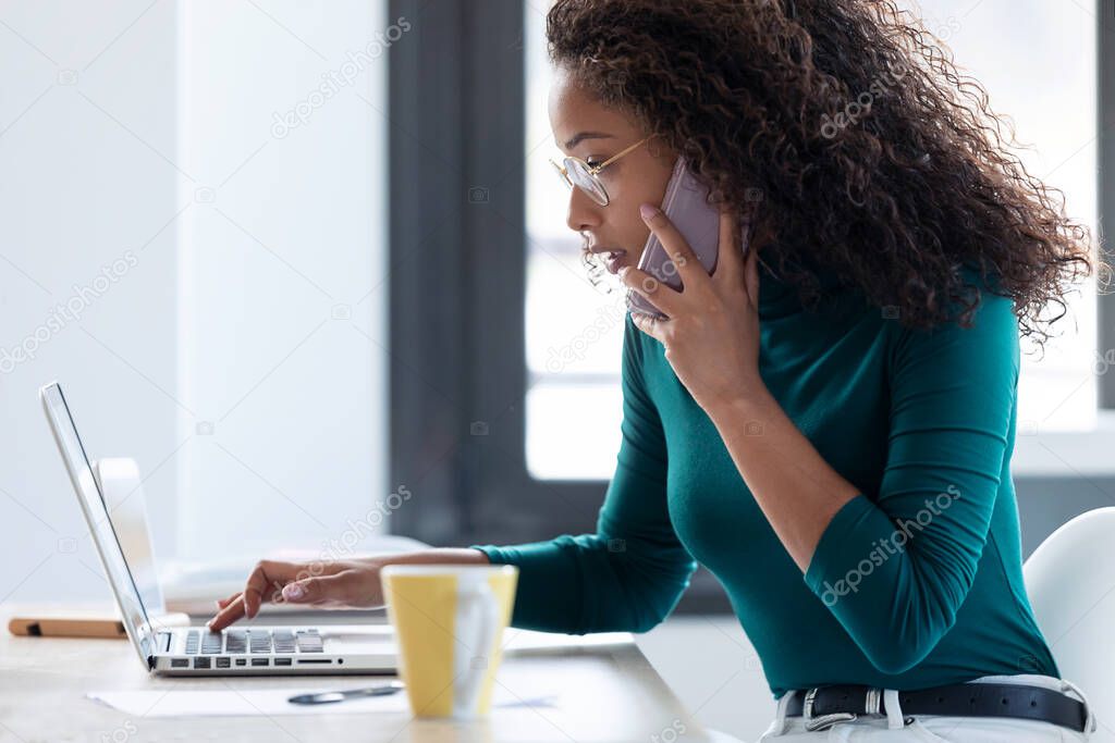 Shot of confident young business woman talking on mobile phone while working with her laptop in the office.