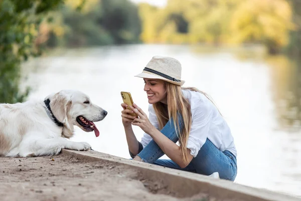 拍了张照片的业余年轻女子坐在公园河边给她的狗拍照 — 图库照片