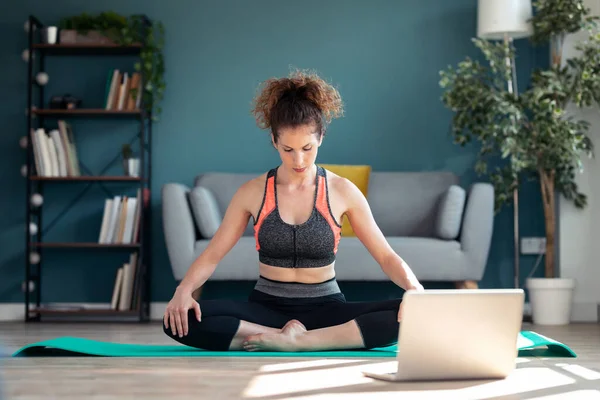 Shot of confident sporty young woman doing hypopressive exercises following online gym classes via laptop on floor in her living room at home.