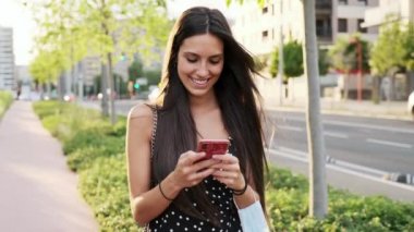 Güzel genç bir kadının koruyucu yüz maskesi tutarken sokakta akıllı telefon ve kısa mesaj kullandığı bir video..