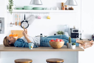 Evdeki mutfaktaki masada uzanırken cep telefonunu kullanan neşeli genç bir kadın..