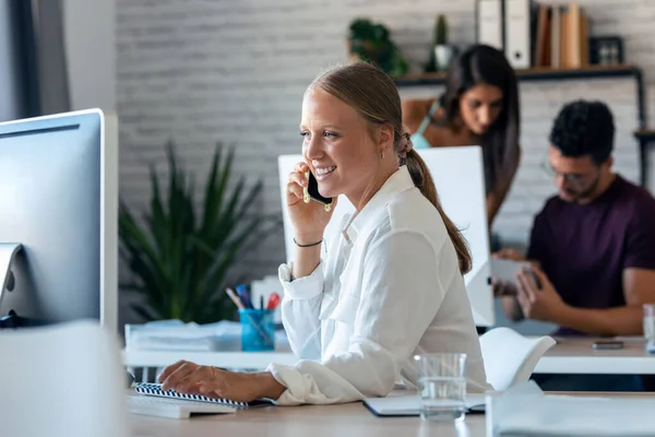 オフィスでパソコンを操作している間に携帯電話で話しているスマートビジネス女性のショット — ストック写真