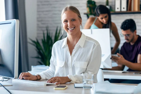 カメラを見ながら笑顔でパソコンを操作している美しい金髪ビジネスの若い女性のショット 背景には 彼の同僚がオフィスで一緒に働いています — ストック写真