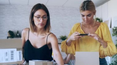 İki güzel serbest çalışan kadının akıllı telefonlarıyla fotoğraf çekerken yeni başladıkları küçük işletmelerde karton kutuları paketleyip mühürledikleri bir video.. 